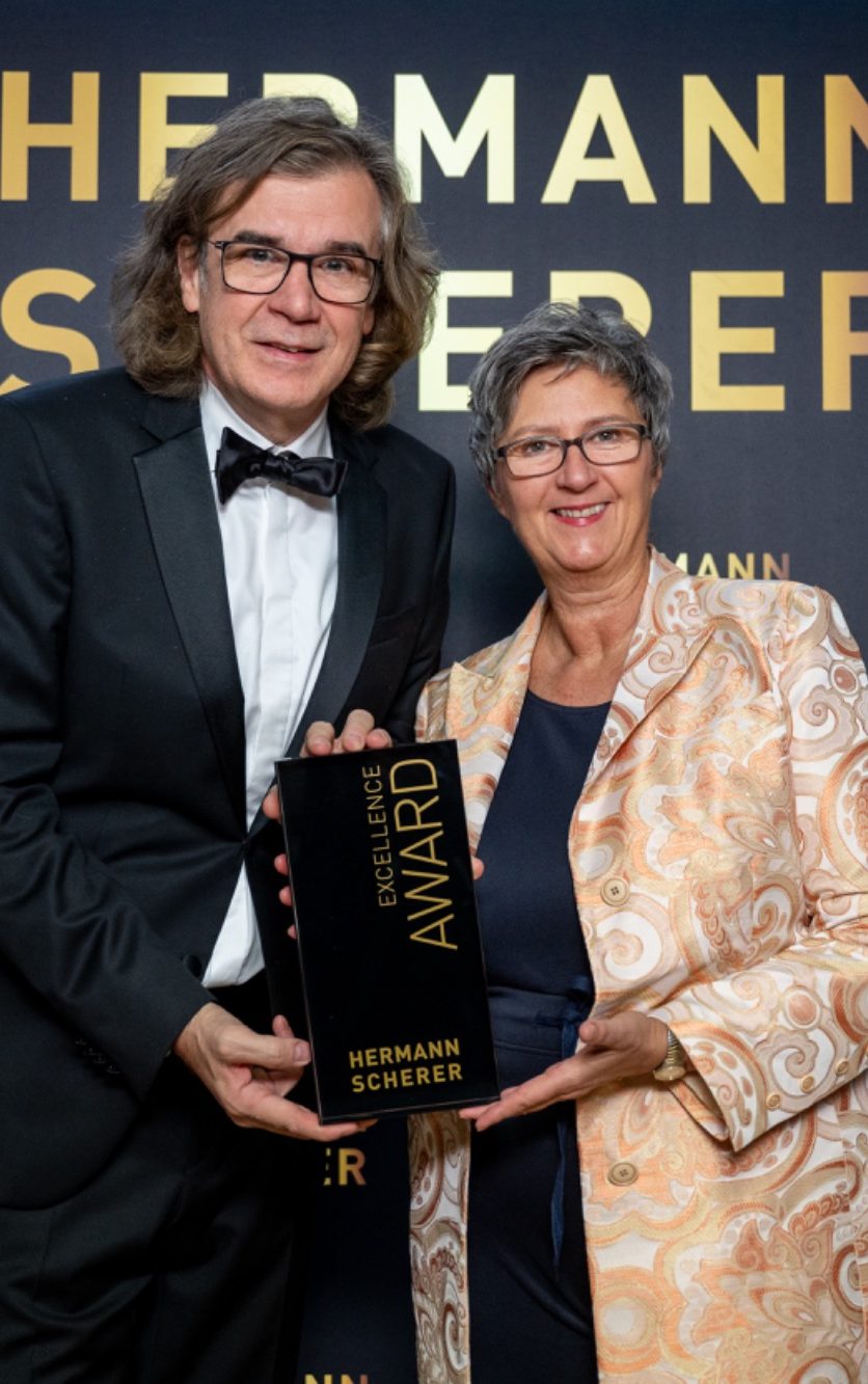 Zwei Personen in formeller Kleidung präsentieren gemeinsam einen Preis auf einer Veranstaltung.