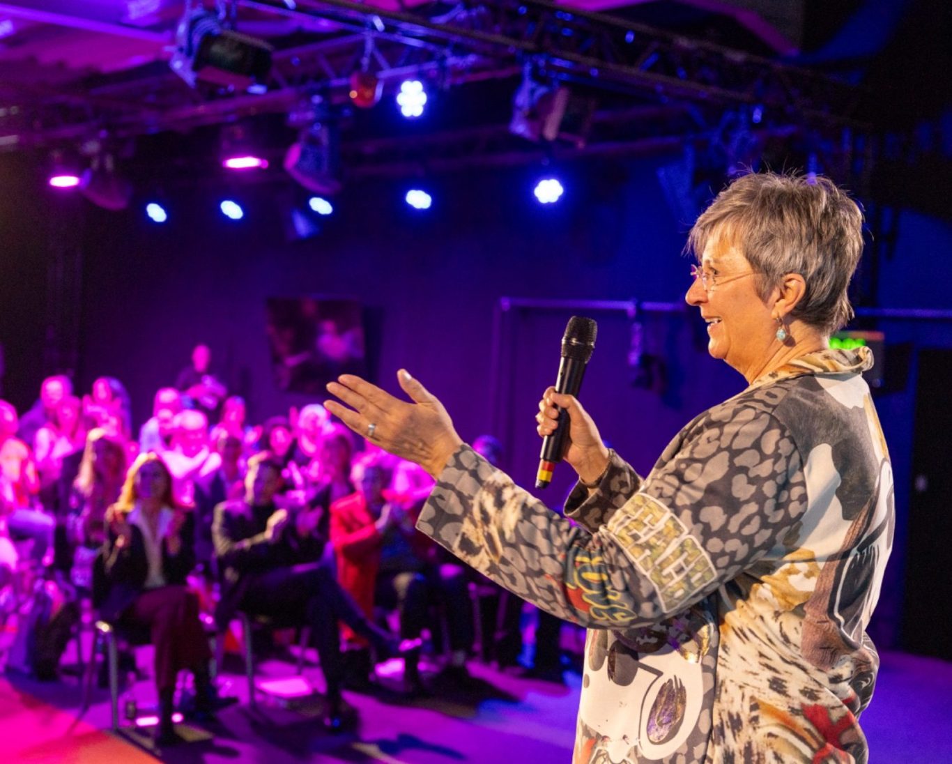 Eine Frau spricht mit einem Mikrofon vor einem Publikum in einem beleuchteten Raum.