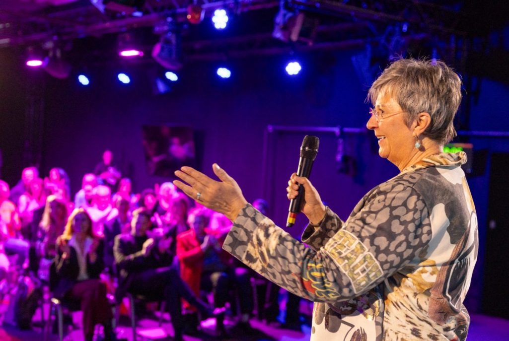 Monika Bröhl im Vortrag Eine Frau spricht mit einem Mikrofon vor einem Publikum in einem beleuchteten Raum.