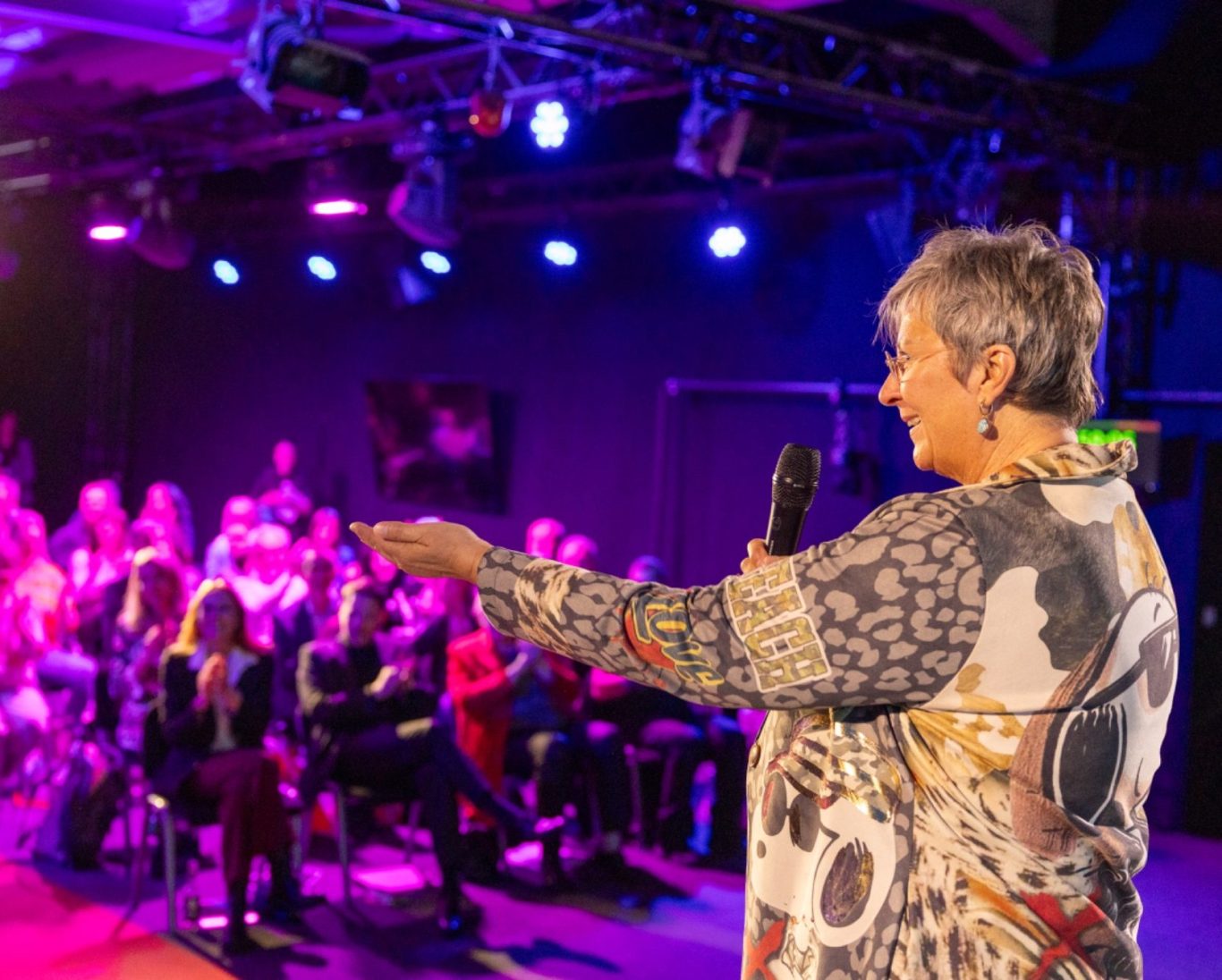 Eine Frau spricht mit einem Mikrofon vor einem Publikum in einem beleuchteten Raum.