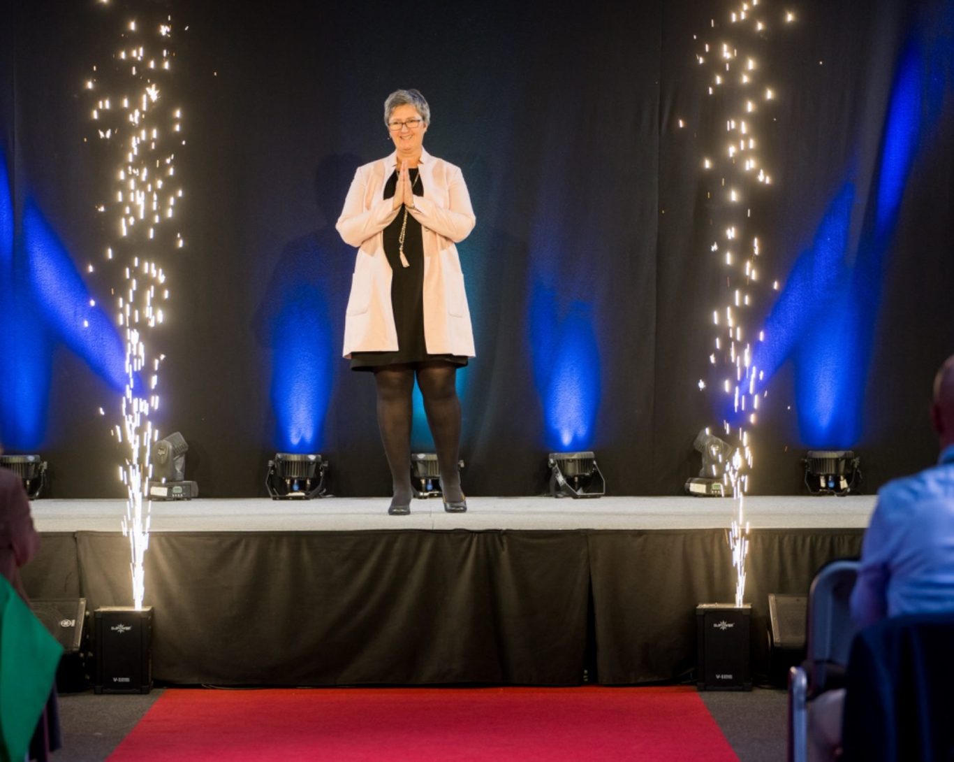 Eine Person in rosa Mantel steht auf einer Bühne, umgeben von Lichtern und rotem Teppich.